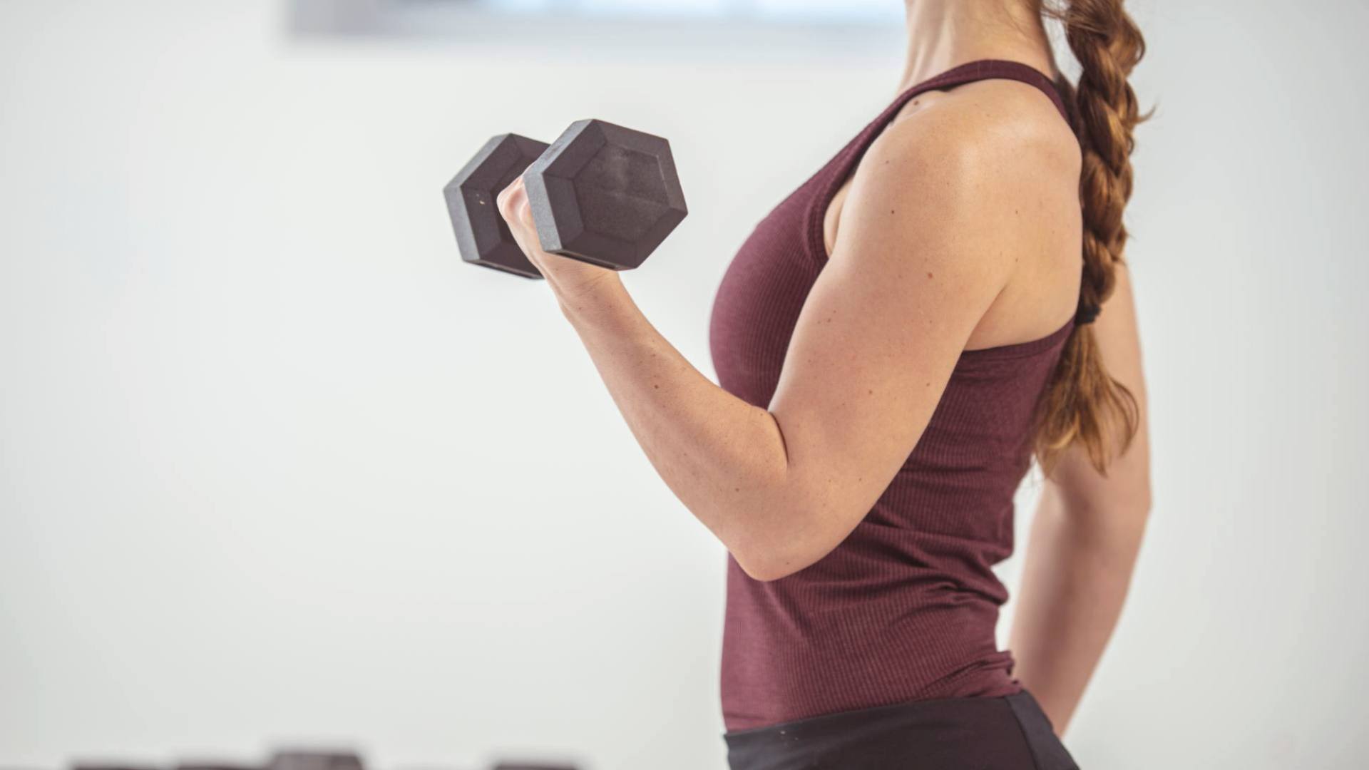 A woman wearing a maroon tank top stands to the side and holds a weight while flexing her left arm, demonstrating the benefits of building muscle.